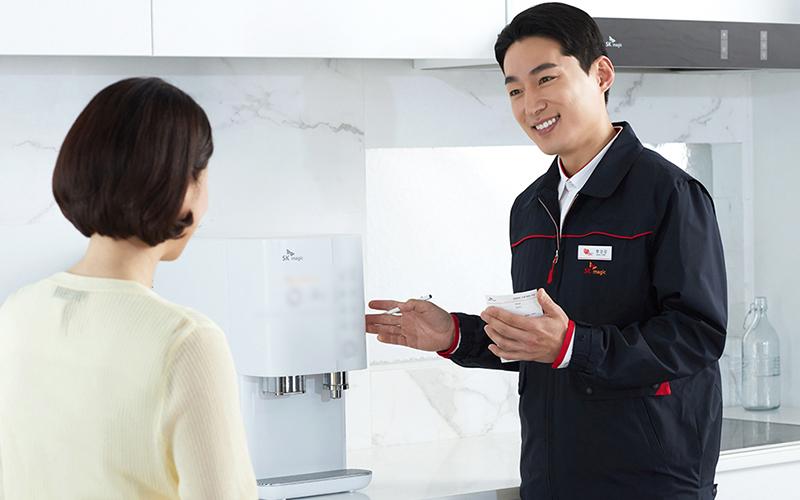 SK매직 정수기 냉수 문제 예방 및 해결 조언 - 검정 유니폼 입은 전문 기사가 흰색 정수기 앞에서 여성 고객에게 친절히 설명하며 손짓하는 장면, 주방 인테리어 배경, 정기 관리·필터 교체·적정 환경 설치 강조